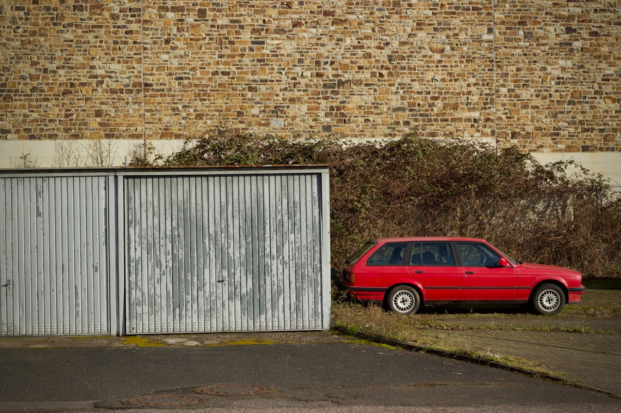 A classic red car parked next to aged garage doors with a rustic brick wall backdrop.